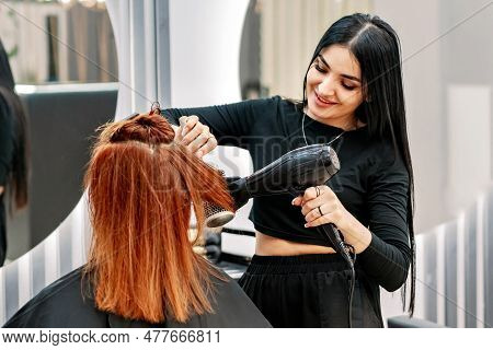 Beautiful Woman Stylist Doing Hair Styling With Hairdryer At Client In Modern Beauty Salon. Hairdres