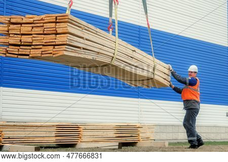 A Slinger In A Helmet And Signal Vest Unloads Wooden Boards On The Street On A Summer Day. Storing B