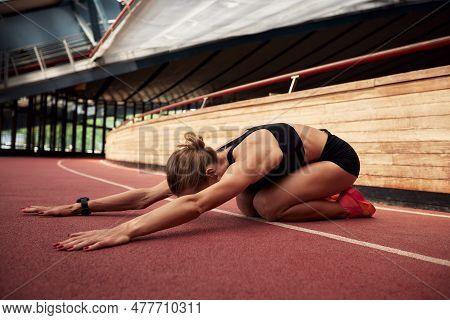 A Young Athlete Girl Is Warming Up Before The Race At The Stadium. A Slender Blonde In A Black Track