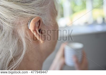 Close Up Of Senior Woman Wearing Behind The Ear Hearing Device Or Aid With Hot Drink At Home
