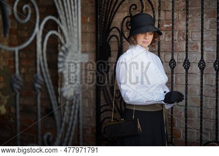 Beautiful Woman In Hat, Gloves, White Blouse And Long Black Skirt, Standing At Black Iron Gate, Bric