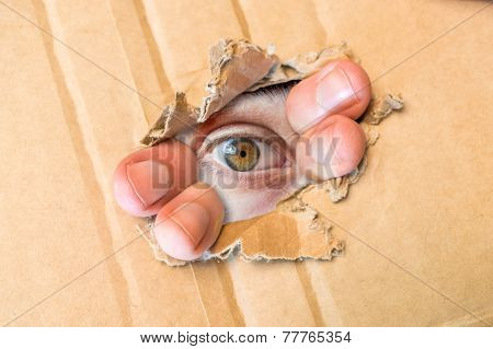 Hidden eye spying through torn hole in cardboard paper