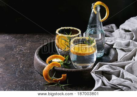 Tonic water cocktail with rosemary and orange. Two glasses and bottle with zest sugar and bubbles in terracotta tray over black texture background. Refreshing beverage alco non alcohol