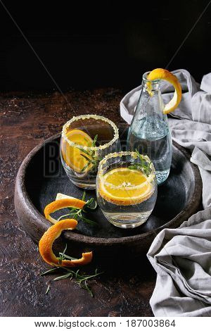 Tonic water cocktail with rosemary and orange. Two glasses and bottle with zest sugar and bubbles in terracotta tray over black texture background. Refreshing beverage alco non alcohol
