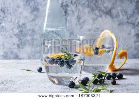 Tonic water cocktail with rosemary, blueberries and orange. Two cold glasses and bottle with zest and bubbles over gray blue texture background. Refreshing beverage alco non alcohol
