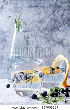 Tonic water cocktail with rosemary, blueberries and orange. Two cold glasses and bottle with zest and bubbles over gray blue texture background. Refreshing beverage alco non alcohol