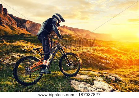 Ryder in full protective equipment on the mtb of two pendant bicycles rides along the green grass towards the sunset in the rays of the sunset sun against the backdrop of a landscape of rocky terrain under the setting sun and clouds.