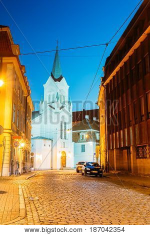 Riga Latvia. The White Tower With Pyramidal Spire Of Our Lady Of Sorrows Or Virgin Of Anguish Church, Ancient Catholic Church On Pils Street In Evening Illumination Under Summer Blue Sky