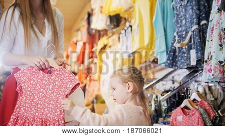 Shopping for kids - a little girl sees a red summer dress with small floral pattern in the mother's arms.