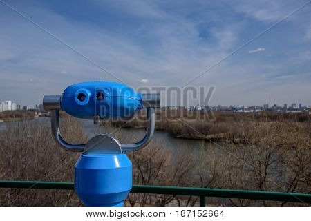 Blue binary telescope of observation deck in park Kolomenskoye, former royal estate, with view of spring forest, Moskva river and the city on the background, Moscow, Russia