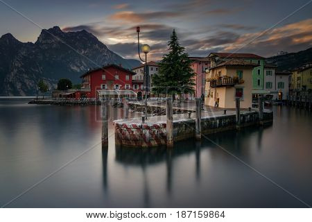 Pier in Torbole harbour at Garda Lake with colorful houses and mountains in background Italy