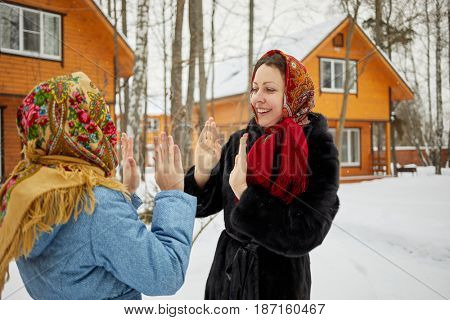 Smiling woman and teenage girl in folk style shawls stand against wooden houses and play okie-dokie on winter day.