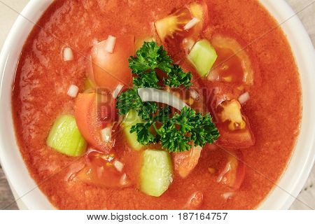 A closeup of a bowl of gazpacho soup with slices of tomato, cucumber, and onion, with parsley leaves