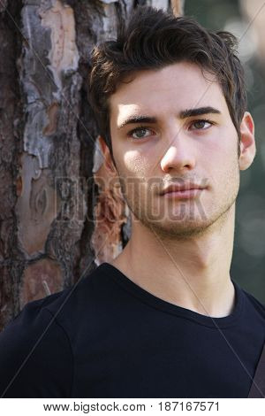 Handsome man close portrait. A beautiful young man with fashionable hair. Looking straight ahead. Behind him a tree. Serene and confident attitude.