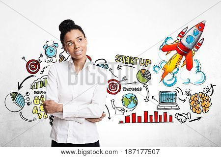 Portrait of an African American businesswoman standing with crossed arms near a concrete wall with a colorful start up sketch and rocket