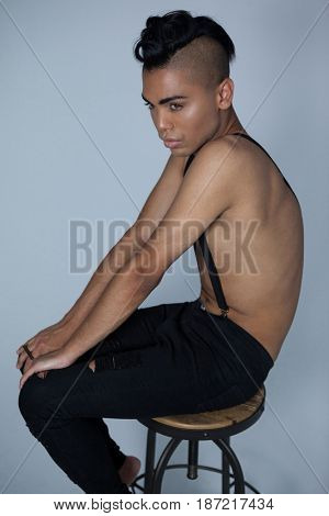 Transgender wearing suspenders sitting on stool against gray background