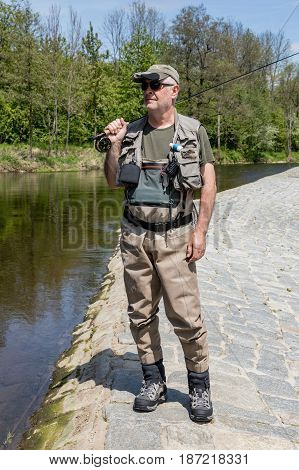 The fisherman walks along the dam on the Otava River. He is dressed in fishing clothes and goes fishing trout. River Otava South Bohemia Czech republic.