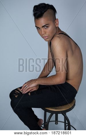 Portrait of transgender wearing suspenders sitting on stool over gray background