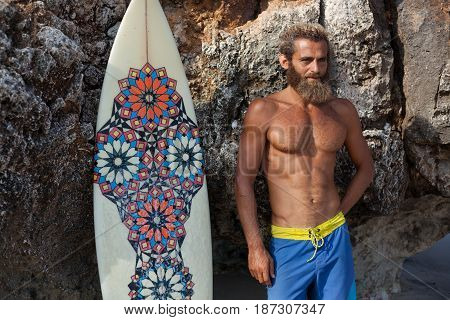Attractive bearded curly man with a surfboard is standing on a beach close to rock