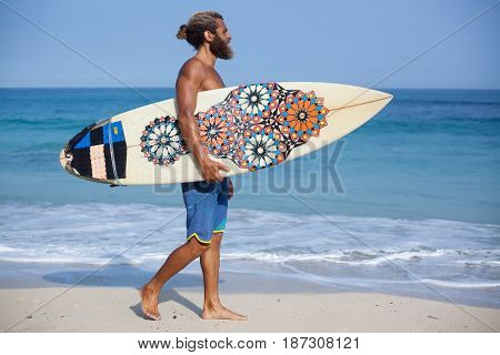 Full-length portrait of an attractive bearded curly man with a surfboard is walking on a beach going from the left side to the right side of the frame