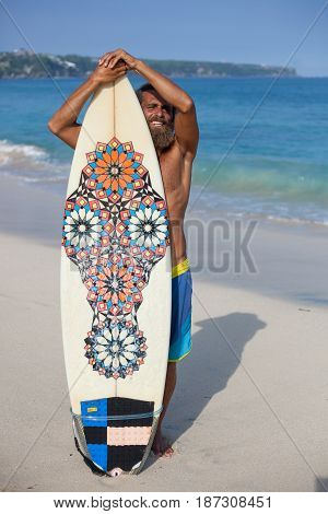 Attractive bearded curly man with surfboard is standing on a beach behind the board and smiling