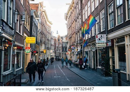 Amsterdam Netherlands - 25 April 2017: Tourist walking along a narrow street of the historical town part of Amsterdam. On the historic houses hang the flags of LGBT.