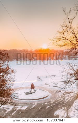Gomel, Belarus. Sun Rising Over City Park At Winter Morning In Gomel, Belarus.  Scenic View Of River Embankment With Statue Of Ancient Man With His Lynx At Sunrise Or Sunset. Lynx - A Symbol Of City.