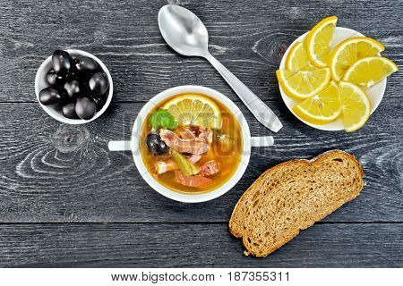 Soup saltwort with lemon, meat, pickles, tomato sauce olives in a bowl, bread and a spoon on the background of a wooden board from above