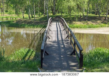 The photo shows a small bridge, a wooden footbridge connecting the island's overgrown island. The footbridge runs over the surface of the lake. It is sunny day.