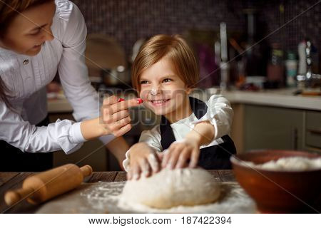 Young woman in formal clothing having fun while making dough with little adorable girl in modern kitchen.
