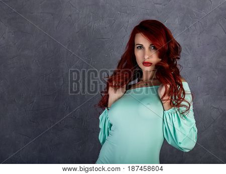 Portrait of beautiful redhead young woman with big boobs in a turquoise dress on a gray background.