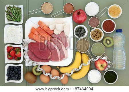 Body building healthy food collection of meat, fish, supplement powders,vitamin pills,fruit, dairy, bottled water and tape measure over slate background.