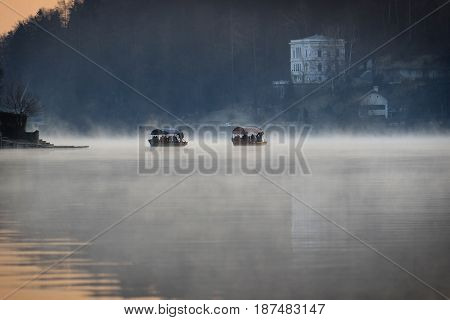 Two pletna boats in Slovenia floaing in mist towards an island inside the lake.