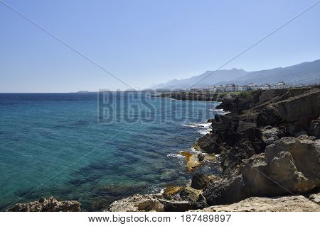Eroded volcano stone at the beach and clear blue water and blue sky with Lapta village in the North of Cyprus in background.
