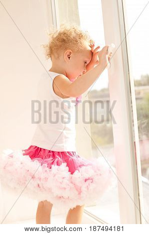 The little girl in a fluffy skirt standing at the open window. A child with white curly hair clings to the window handle. The little girl looks out the window.