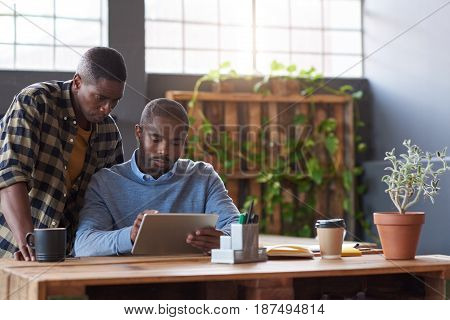 Two casually dressed young African business colleagues using a digital tablet and talking together while working at a table in a modern office