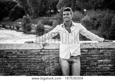 Handsome smiling man outdoors. Tiber river from the bridge. A beautiful young Italian man relaxing on Milvian Bridge in the historic center of Rome, Italy. Blue shirt and jeans. Black and white.