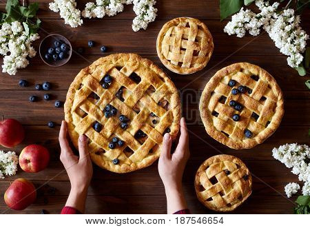 Homemade organic apple pie pies ready to eat hold female hands on dark wooden table with flowers, blueberry and apples. Traditional dessert on Independence Day. Food flat lay background