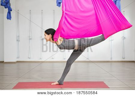 Girl stretching leg with help of hammock. Aerial exercise or antigravity yoga indoors, woman does aero poses with head leaned to back
