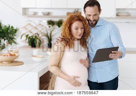 Family moments. Mature husband embracing his pregnant wife and showing her some videos on a table while she is sitting on a bar stool.
