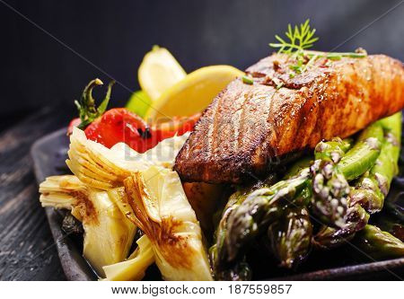 Recipe - delicious portion of grilled salmon fillet with asparagus tomatoes artichoke spices on a black wooden board. Close up copyspace selective focus.