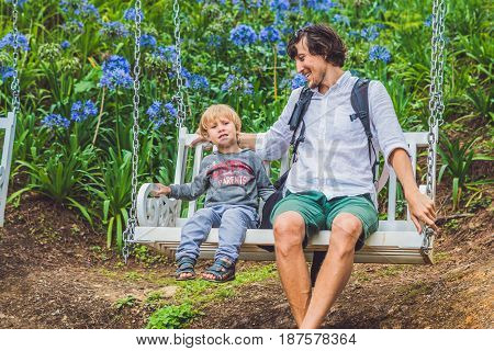 Father And Son Having Fun On Swings In A Flower Garden