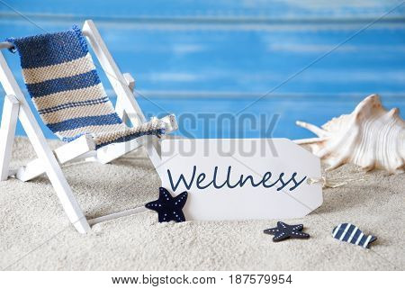 Summer Label With English Text Wellness. Blue Wooden Background. Card With Holiday Greetings. Beach Vacation Symbolized By Sand, Deck Chair And Shell.