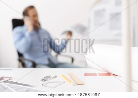 Tools for work. Clse up of drawing tools lying on the table while professional senior engineer talking on smart phone in the background