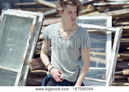 Portrait of young handsome man over wood boards, outdoor. Attractive trendy fashion 18 years old teen boy posing outside over debris, image toned