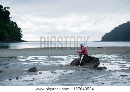 Woman Sitting On Rock Among Moist Sand Beach. Front Of Her Have Blue Sea,sky And Green Forest On Mou