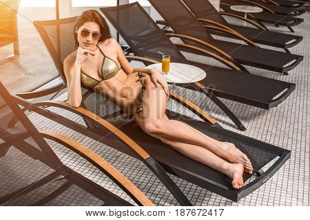 Young attractive slim girl in bikini relaxing on deck chair in wellness spa hotel resort. Near the table is a glass of orange juice. sun flare