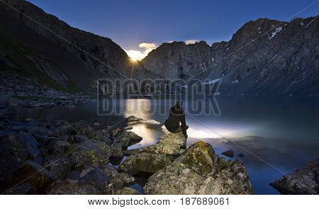 man in black jacket with headlight sitting near blue lake Alakol in mountains at night with mountains surrounded and moon rising