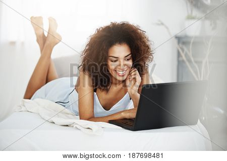 Young happy beautiful african girl in sleepwear lying on bed at home smiling looking at laptop.