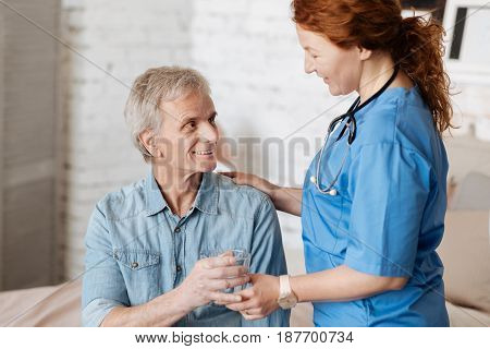 Refreshing drink. Charismatic motivated mindful woman making sure her patient feeling better while serving him a glass of water during the regular visiting at home